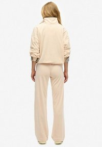 Vrouw met lang blond haar, gekleed in losse beige broek en bijpassend langemouwen top, staand tegen een effen witte achtergrond.