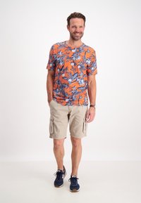 Camicetta a maniche corte con motivo floreale arancione e dettagli blu, abbinata a pantaloni cargo beige e sneaker blu navy. Vestibilità casual e materiale leggero.