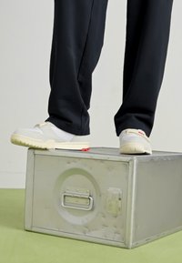 Sneakers bianchi con tomaia testurizzata, suola beige e accento rosso, posizionati su una scatola metallica, con pantaloni neri in un ambiente minimalista.
