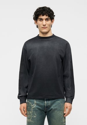 Jeune homme aux cheveux bouclés portant une chemise noire à manches longues et un jean bleu avec des motifs graphiques, debout devant un fond clair uni.