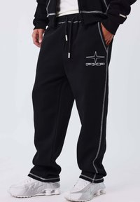 Hombre vestido con pantalones deportivos negros con costuras blancas y logo de PXP, combinado con zapatillas Nike blancas, de pie frente a un fondo liso.