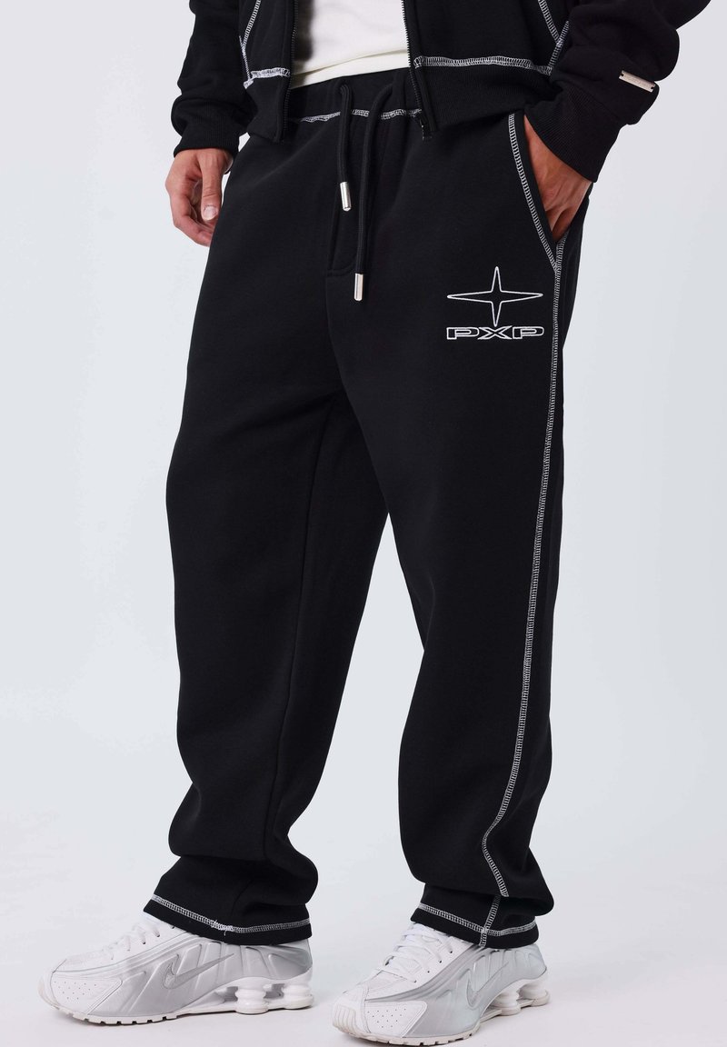 Hombre vestido con pantalones deportivos negros con costuras blancas y logo de PXP, combinado con zapatillas Nike blancas, de pie frente a un fondo liso.