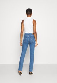Hoch geschnittene blaue Denim-Jeans verfügen über ein klassisches Fünf-Taschen-Design und charakteristische Rückennähte. Kombiniert mit einem ärmellosen weißen Oberteil.