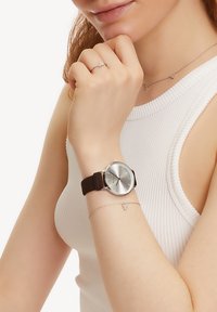 Braune Silikonarmbanduhr mit einem runden silbernen Zifferblatt, das silberne Zeiger und ein einfaches Design aufweist. Zarte Silberkettengliederarmband mit Herzdetails.