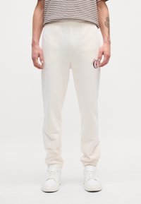 Kremowe spodnie dresowe z elastycznym pasem, zwężanymi nogawkami i subtelnym logo "Calvin Klein" w burgundowym kolorze na lewej udzie. Noszone z białymi sneakersami.