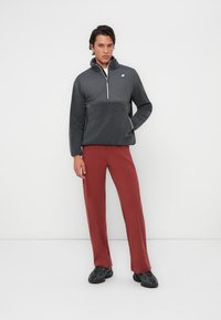 Pullover in donkergrijze fleece met een rits hals, gecombineerd met tapered roestkleurige broek. Zwarte sportschoenen maken de outfit compleet.