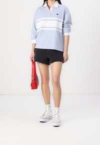 Polo azzurro chiaro a maniche lunghe con strisce bianche, pantaloni corti di jeans neri, sneakers bianche alte e una piccola borsa rossa.