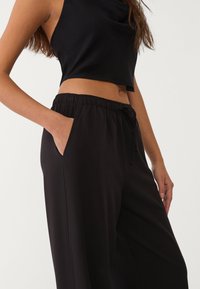 Schwarzes Cropped-Top mit drapierter Gestaltung, kombiniert mit lockeren schwarzen Hosen. Die Hosen verfügen über einen elastischen Bund und seitliche Taschen.
