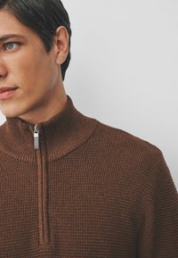 Brauner Strickpullover mit hohem Kragen und Viertelreißverschluss. Das strukturierte Muster und die gerippten Bündchen verleihen dem Design Details.