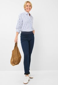 Camicia a righe blu e bianche, jeans blu scuro, sneakers bianche e una borsa in suede marrone con borchie metalliche. Vestibilità rilassata, maniche arrotolate, stile casual.
