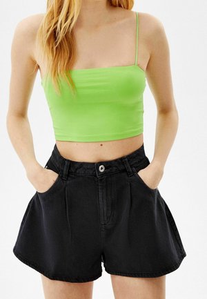 Femme portant un crop top vert citron à fines bretelles et un short noir taille haute à jambes larges, les mains dans les poches.