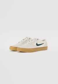 Sportovní boty v béžové semišové kůži s bílými akcenty a tmavozeleným logem ve tvaru klíče. Gumová podrážka, plochý design a kulaté tkaničky.