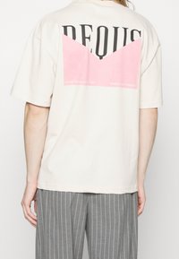 Jasnoniebieska, oversize koszulka z krótkim rękawem, z różowym geometrycznym wzorem na plecach oraz wyraźnym czarnym napisem.