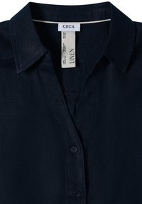 Chemise en lin bleu marine foncé avec col et boutonnage devant, avec une étiquette blanche portant la marque "CECIL" et le texte "LINEN".