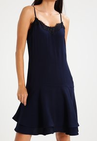 Marinblå slipsklänning med spetskant, med tunna justerbara band och en utsvängd nederdel. Tillverkad av ett slätt material med en subtil lyster.