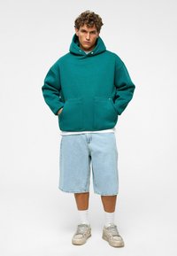 Teal oversized hoodie met kangoeroezakken, gemaakt van een katoenmix, gecombineerd met lichtblauwe denim shorts en grijze sneakers. Effen achtergrond.