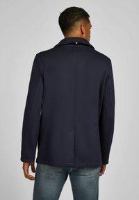 Dunkelblaues Jackett mit strukturiertem Aussehen, ausgestattet mit einem spitzen Kragen und seitlichen Taschen. Der Stoff ist glatt und hat eine körpernahe Passform.