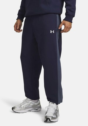 Man die marineblauwe Under Armour joggingbroek en zilverkleurige sportschoenen draagt, staand tegen een effen achtergrond.