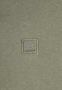 Groene stof met een gestructureerd breipatroon en een vierkant, gestikt logo-patch in het midden met het label "GABBIAINO".