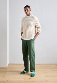 Maglione a maglia crema con texture a coste, pantaloni di cotone verdi e sneakers verdi con strisce bianche. Posizione in piedi su uno sfondo chiaro.
