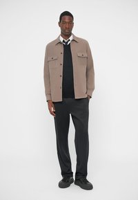 Bezs színű gombos dzseki két mellzsebbel, fekete pulóver és fekete nadrág felett viselve. A modell áll, fekete cipőt visel.