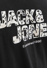 Sort bomulds T-shirt med fed, hvid camouflage-mønster tekst "JACK & JONES" med "Established 1990" i mindre tryk nedenfor.