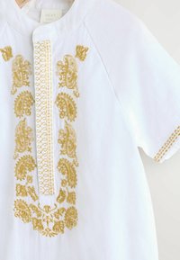 Chemise en lin blanc avec une broderie dorée complexe sur le devant et l'encolure, manches courtes et surpiqûres décoratives aux poignets.