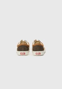 Pantofi casual maro și bej din materiale de nubuc și piele, cu tălpi din cauciuc alb și un logo roșu pe călcâi. Au un design de tip low-top.