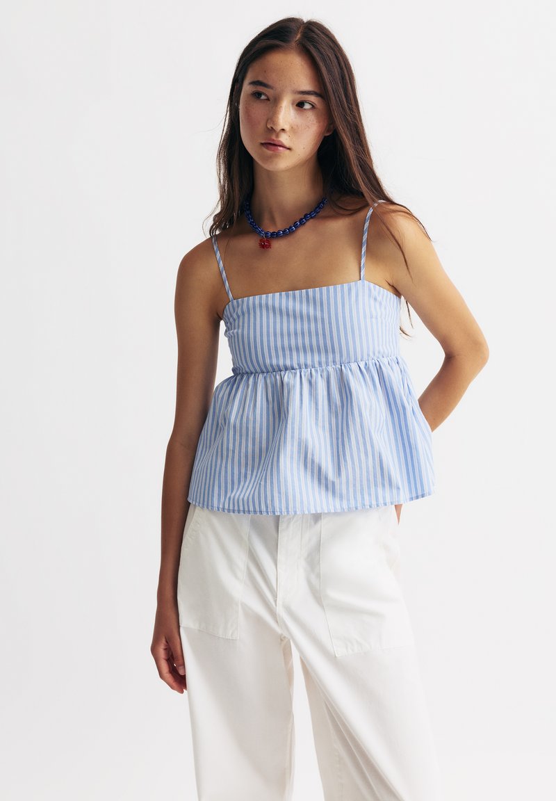 Modrý a bílý pruhovaný top s peplum střihem, tenkými ramínky a nařaseným pasem, spárovaný s bílými kalhotami s vysokým pasem a modrým náhrdelníkem.