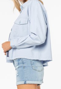 Světle modrá džínsová bunda s dlouhými rukávy, zapínání na knoflíky, dvě přední kapsy a třepený lem. V kombinaci se světle modrými džínovými šortkami s obnošeným vzhledem.