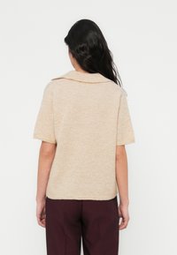 Πουλόβερ με κοντά μανίκια, μπεζ πλεκτό με γιακά. Μαλακή υφή, διακριτικές αποχρώσεις στο χρώμα. Φορεμένο με σκούρο βυσσινί παντελόνι.