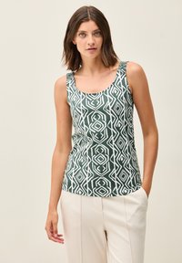 Ärmös grön linne med ett vitt geometriskt mönster, gjort av ett mjukt tyg. Rundad nacke, figursydd form. Bärs med beige byxor.