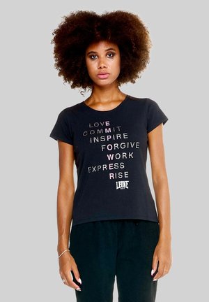 Femme aux cheveux bouclés portant un t-shirt noir avec des mots motivants et "Empower" écrit verticalement en lettres roses.