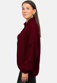 Pull en tricot bordeaux avec col montant, coupe oversize et textures côtelées. Manches longues bouffantes et fentes latérales à l'ourlet.