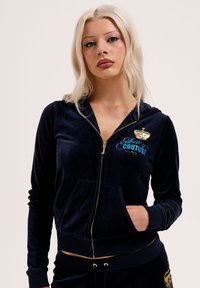 Femme aux longs cheveux blonds portant un sweat à capuche bleu marine en velours zippé et un pantalon assorti avec une couronne dorée et une broderie "Juicy Couture" bleue.