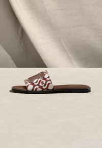 Επίπεδο σανδάλι με καφέ σόλα και φαρδιά λουρί με κυματιστό κόκκινο και λευκό μοτίβο και καφέ δερμάτινο ταμπελάκι με λογότυπο.