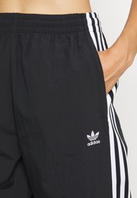 Černé sportovní kalhoty s elastickým pasem, bílými bočními pruhy a malým bílým logem Adidas na levém boku. Hladká textura.