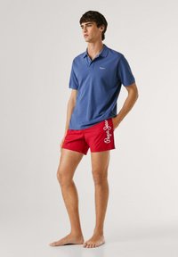 Jaunas vyras basas stovintis, dėvintis mėlyną polo marškinėlį ir raudonus maudymosi šortus su baltu užrašu „Pepe Jeans“ kairėje kojoje.