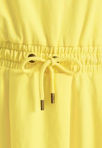 Gult tyg med en slät yta och rynkad elastisk midja. Har en dragsko med guldfärgade metalldelar och matchande öljett. 