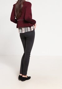 Cardigan texturé bordeaux porté sur un chemisier rayé, avec un jean noir slim et des chaussures noires, vu de dos.
