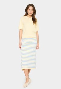 Polo shirt jaune à manches courtes, texture lisse. Jupe rayée, longueur genou, en bleu clair et crème ; tissu léger. Chaussures espadrilles soigneusement nouées.