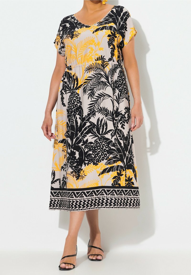 Femme portant une robe midi à manches courtes, avec un motif de feuilles tropicales et ananas noir et jaune sur un fond clair.