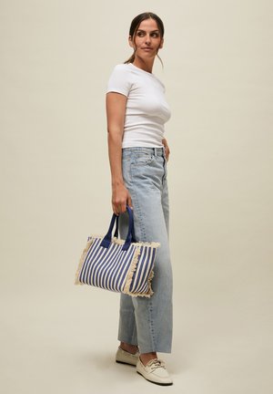 Vrouw draagt wit T-shirt en lichtblauwe spijkerbroek, houdt een blauw-wit gestreepte tas met franjes vast, staand tegen een beige achtergrond.