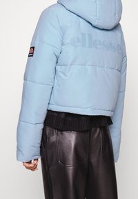 Jasnoniebieska krótka kurtka puchowa z kapturem, z subtelnym nadrukiem logo na plecach i czarnymi prążkowanymi detalami u dołu. Czarne skórzane spodnie.