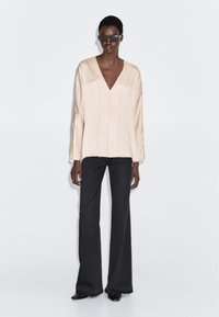 Beige long-sleeve blouse with a deep V-neck and vertical seams, paired with high-waisted black flared jeans. Model wearing dark sunglasses.