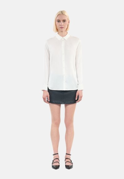 Witte blouse met lange mouwen en knoopsluiting, gecombineerd met een zwarte leren mini-jurk en zwarte sandalen met bandjes. Soepele stoftextuur.