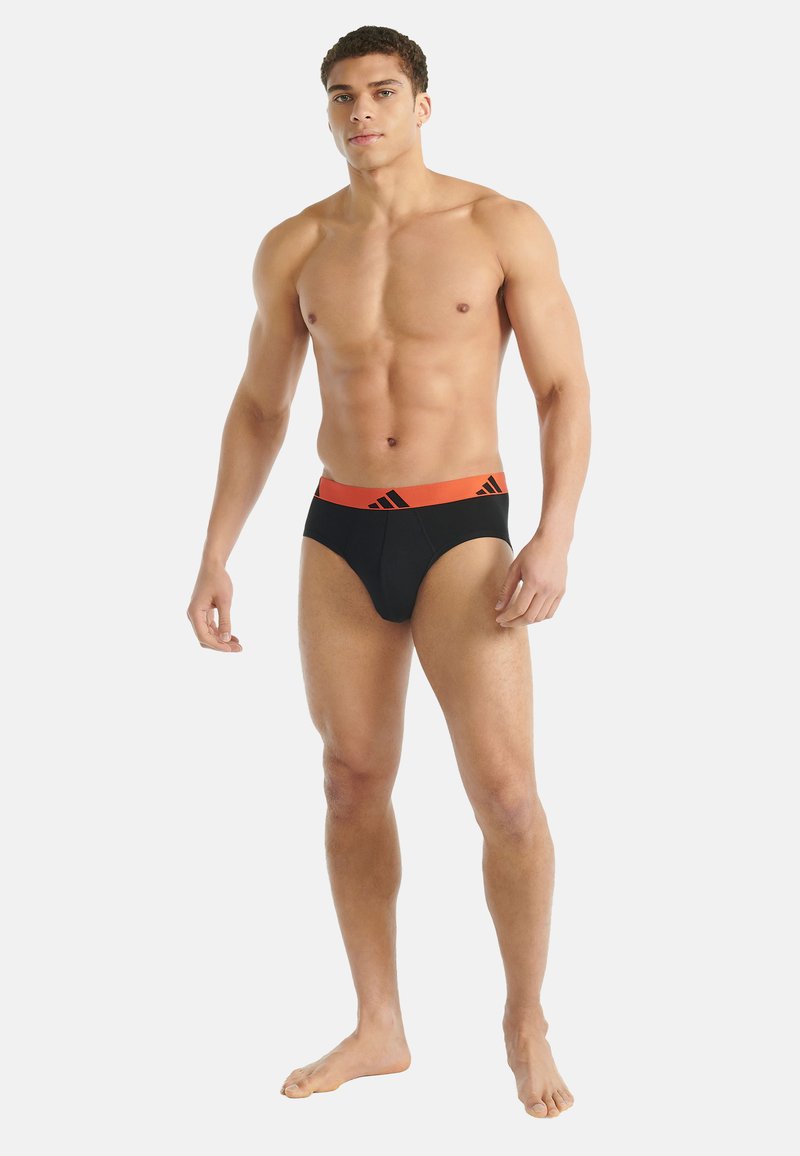Czarne męskie slipy z pomarańczową gumką w pasie z logo. Tkanina wydaje się gładka, a dopasowanie jest przylegające, podkreślając kształt ciała.