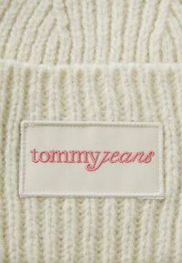 Kreemivärviline kudum, millel on ribilised tekstuurid. Sisaldab ristküliku kujulist sildit, millel on roosa kalligraafia "tommyjeans".