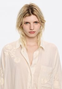 Camicia button-up beige chiaro con ricami floreali sulla spalla sinistra e sulla tasca; tessuto morbido e texture con vestibilità rilassata.