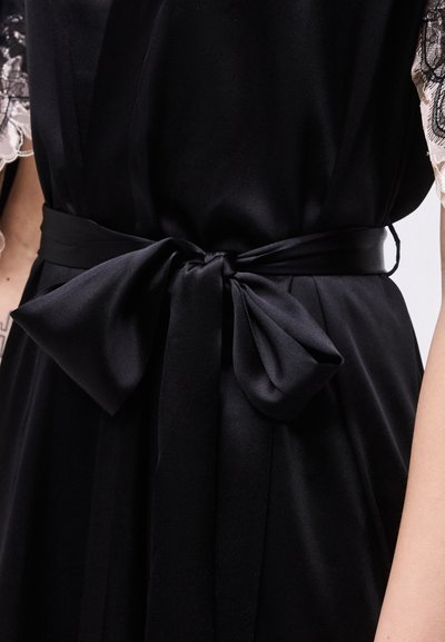 Μαύρο σατέν φόρεμα με δαντελωτά μανίκια, με φαρδύ ζωνάρι που δένεται σε μεγάλο φιόγκο στη μέση.